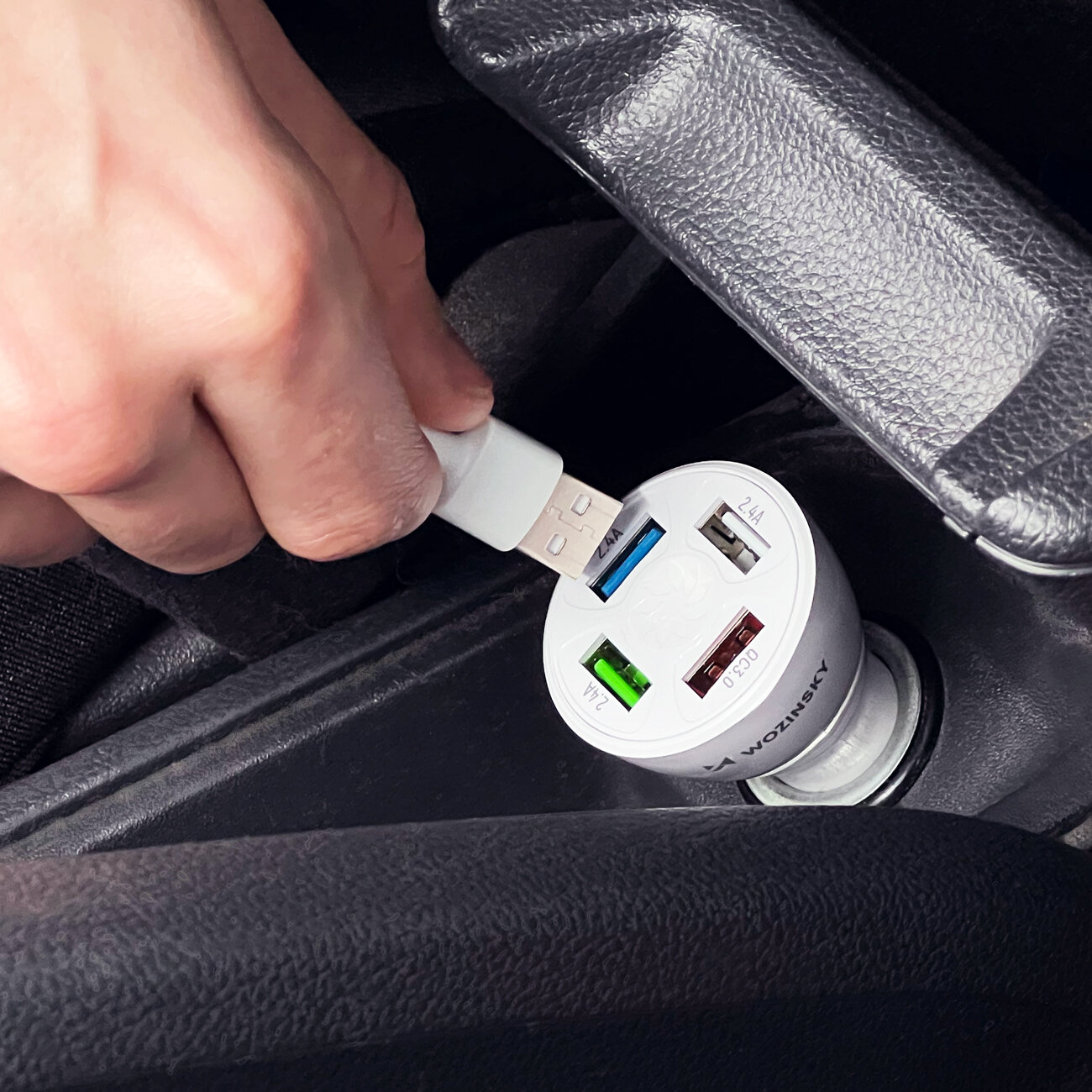 Osoba wkłada kabel USB do białej ładowarki samochodowej z czterema portami, umieszczonej w gnieździe zapalniczki. Na ładowarce widoczne są oznaczenia portów oraz napis \'Wozinsky\'.