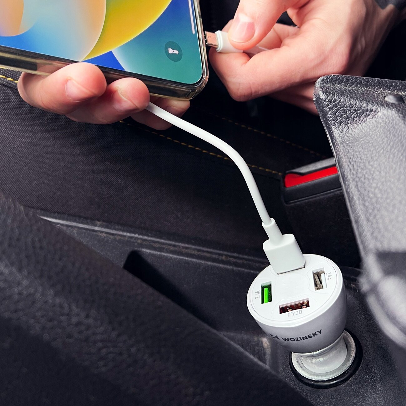 Osoba podłącza kabel do telefonu, który jest połączony z ładowarką samochodową w gnieździe zapalniczki. Na ładowarce widoczne są cztery porty USB i napis \'Wozinsky\'.