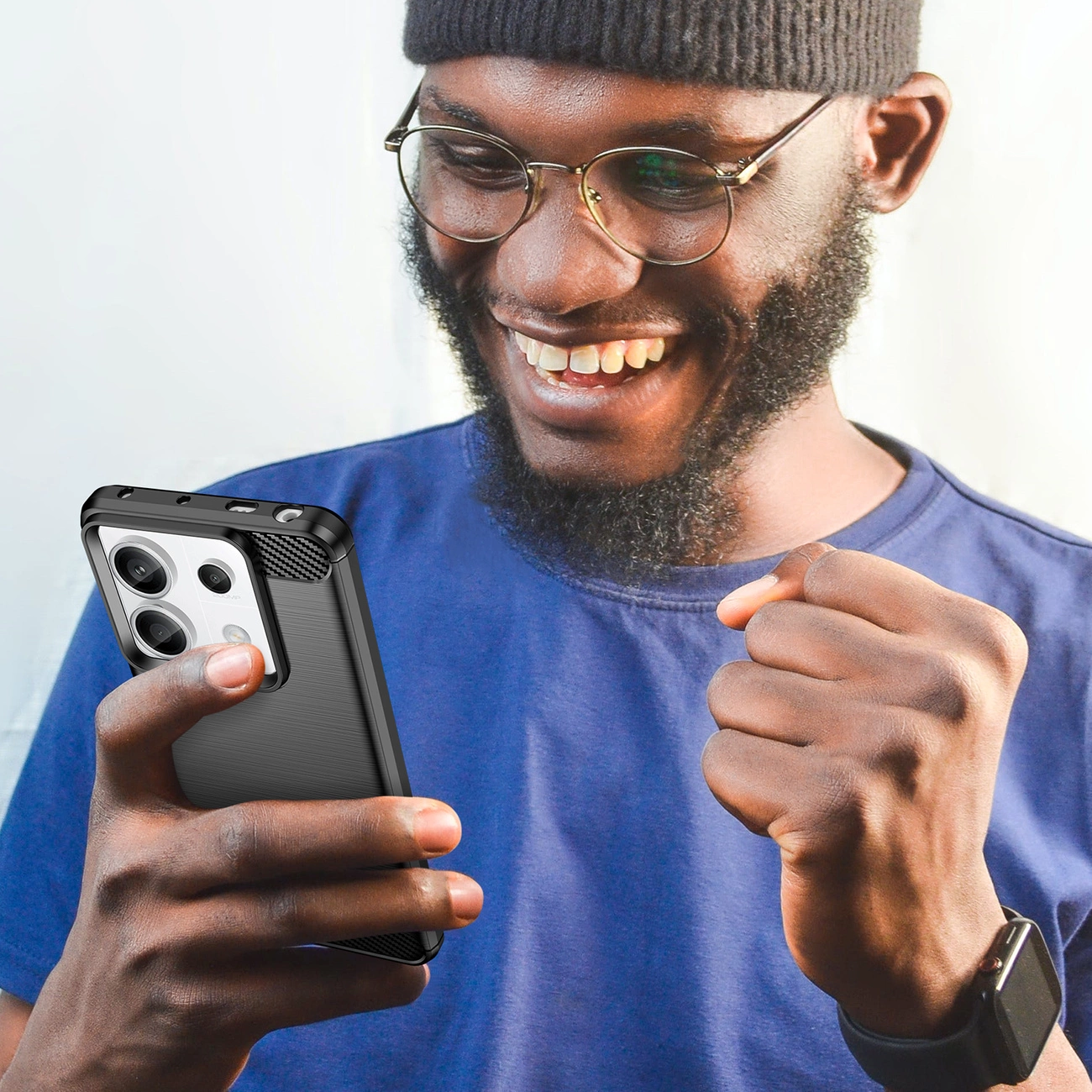 Osoba trzyma smartfon w czarnym etui z widocznymi trzema obiektywami aparatu. Osoba wygląda na zadowoloną, ma na sobie niebieską koszulkę i czarną czapkę.
