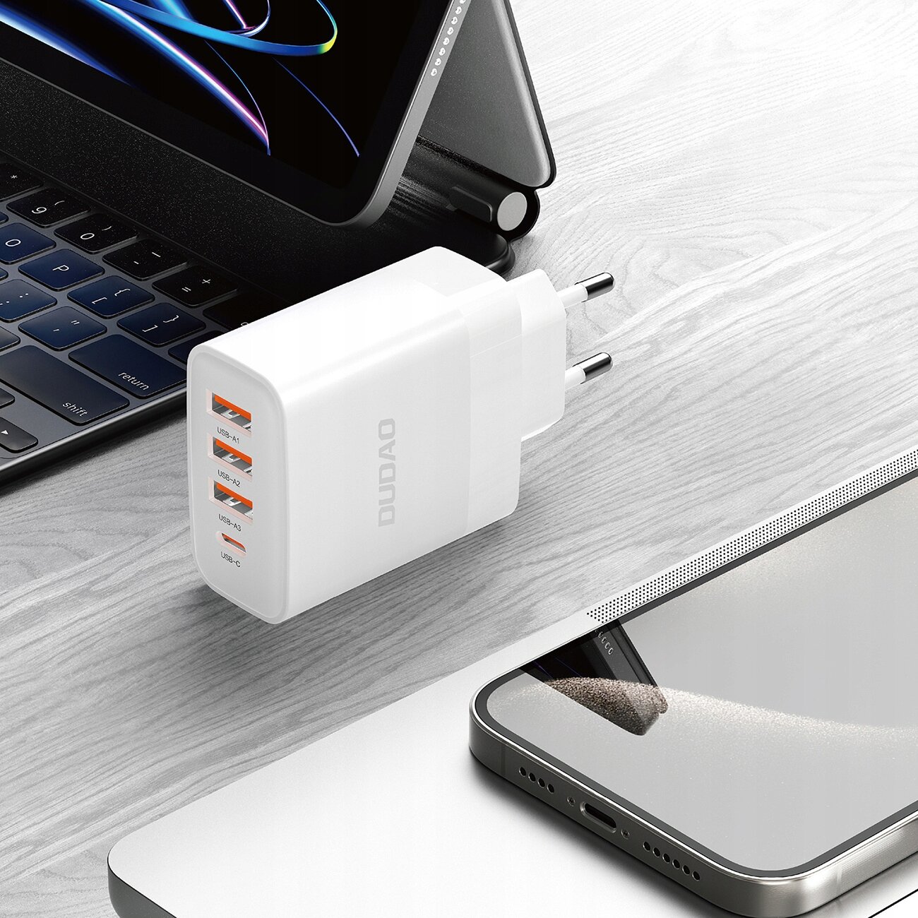 Na drewnianym blacie znajduje się biała ładowarka z logo \'DUDAO\', posiadająca trzy porty USB-A i jeden port USB-C. Obok widoczne są fragmenty laptopa i smartfona.