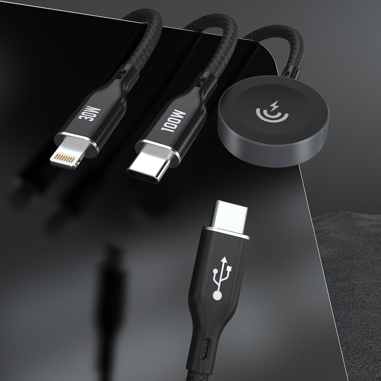 Na obrazie widoczne są trzy czarne kable z różnymi końcówkami: Lightning z oznaczeniem \'30W\', USB-C z oznaczeniem \'100W\' oraz okrągła końcówka z symbolem ładowania. Kable leżą na ciemnej powierzchni, a jeden z nich ma symbol USB na wtyczce.