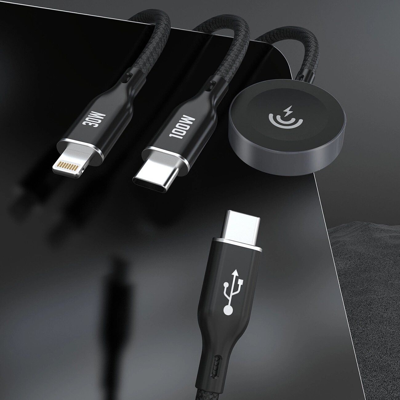 Na obrazie widoczne są trzy czarne kable z różnymi końcówkami: Lightning z oznaczeniem 30W, USB-C z oznaczeniem 100W oraz okrągła końcówka z symbolem ładowania. Kable leżą na ciemnej powierzchni.
