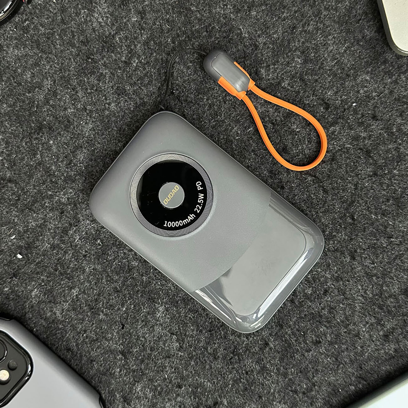 Na obrazie widoczny jest szary powerbank z napisem \'DUDAO 10000mAh 22.5W PD\' na okrągłym czarnym elemencie. Obok znajduje się pomarańczowy kabel z szarymi końcówkami.