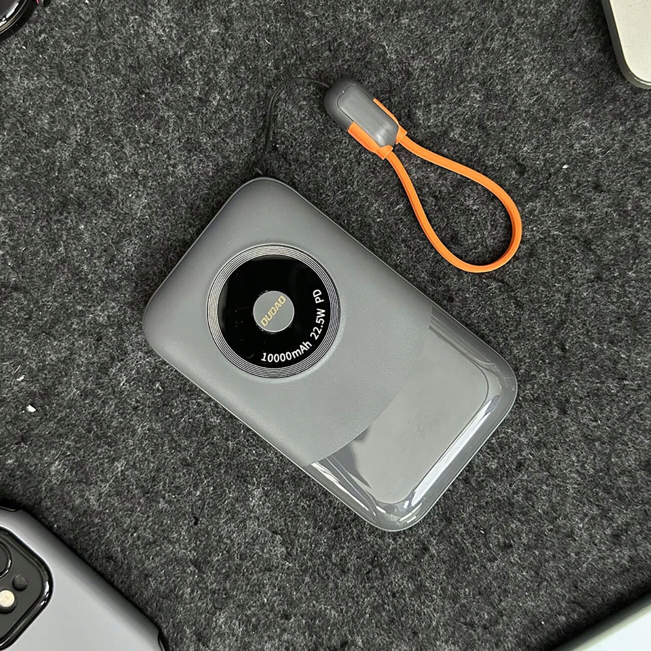 Na szarej powierzchni leży szary powerbank z okrągłym wyświetlaczem, na którym widnieje napis \'DUDAO 10000mAh 22.5W PD\'. Obok znajduje się pomarańczowy kabel z szarymi końcówkami.