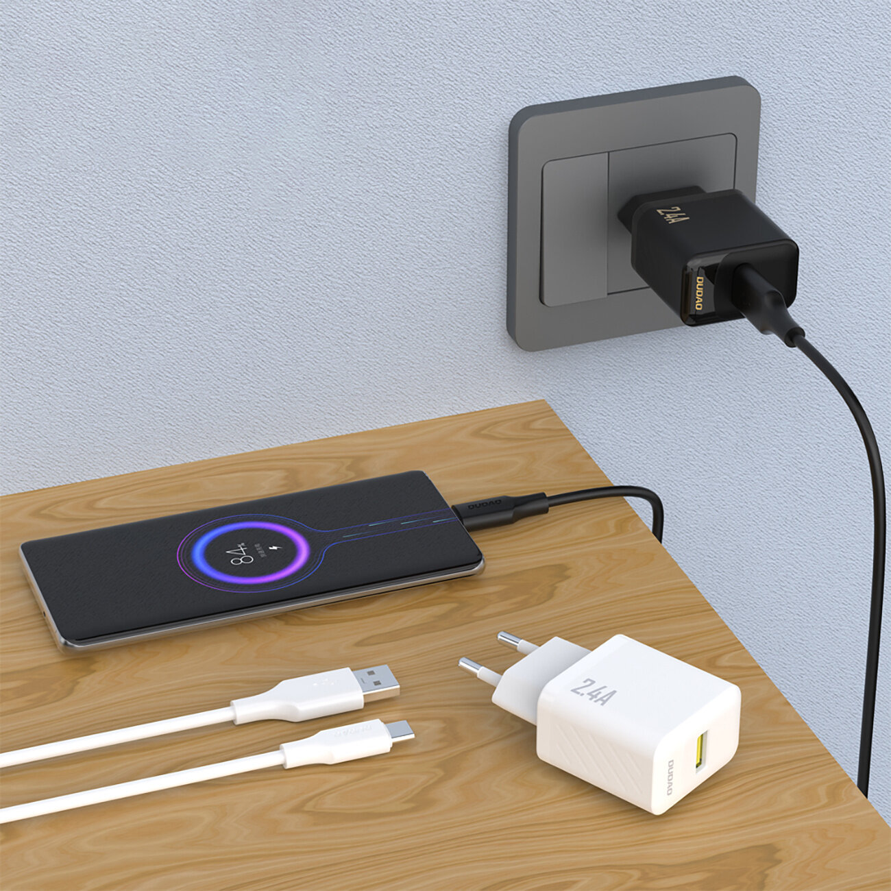 Na drewnianym blacie leży biały adapter z napisem \'2.4A\' oraz dwa kable USB. W tle widać telefon podłączony do czarnej ładowarki w gniazdku ściennym.