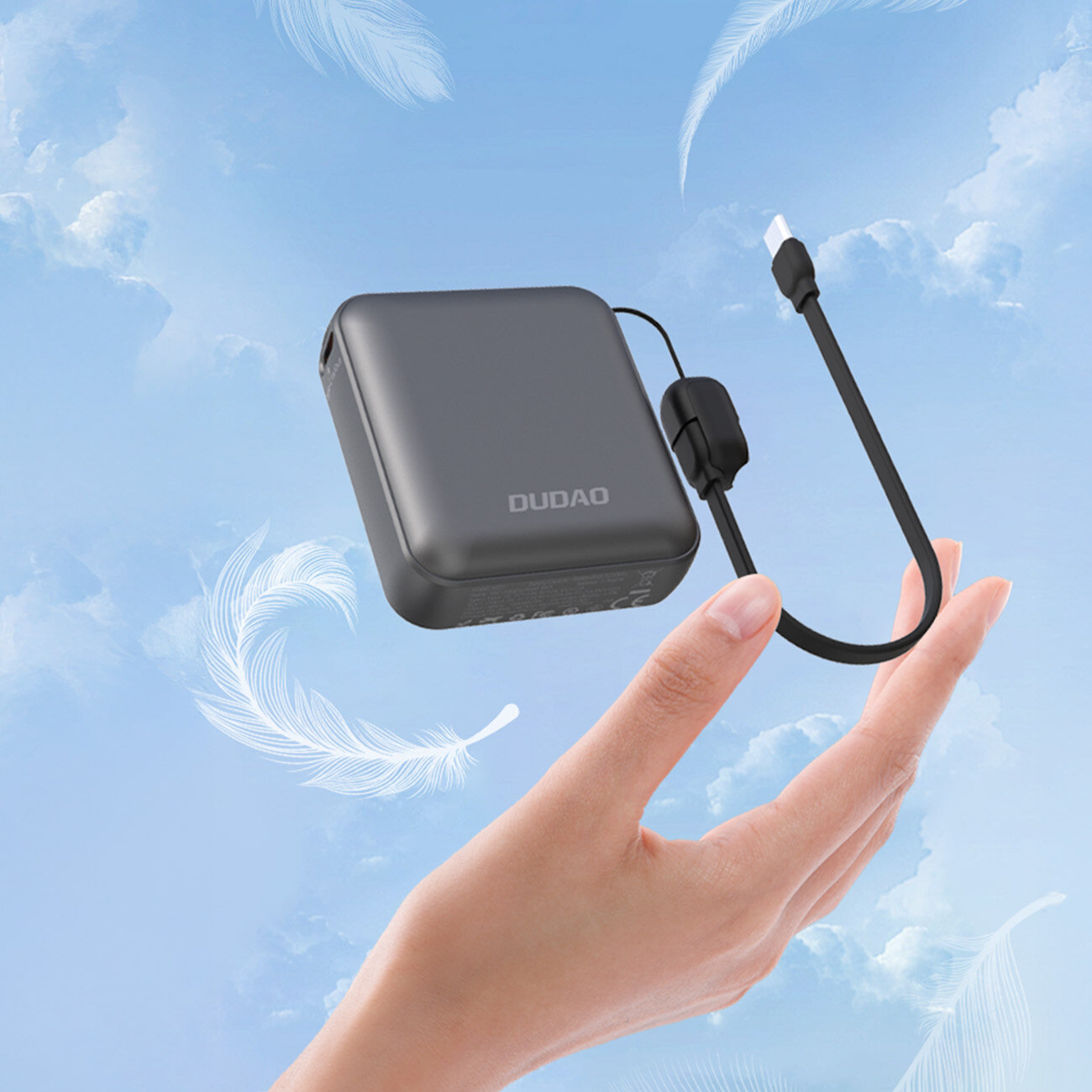 Na tle nieba z piórami unosi się szary powerbank z napisem \'DUDAO\', trzymany przez dłoń. Do urządzenia podłączony jest czarny kabel USB-C.