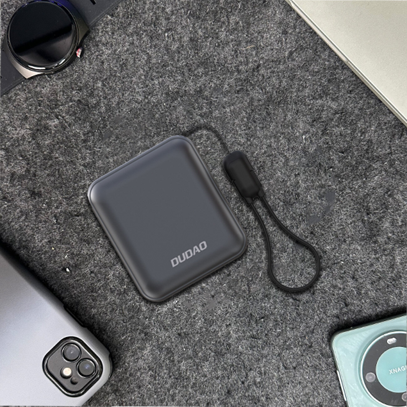 Na szarej powierzchni leży szary powerbank z napisem \'DUDAO\' oraz czarną zawieszką. Wokół znajdują się różne urządzenia elektroniczne, w tym smartfon i zegarek.