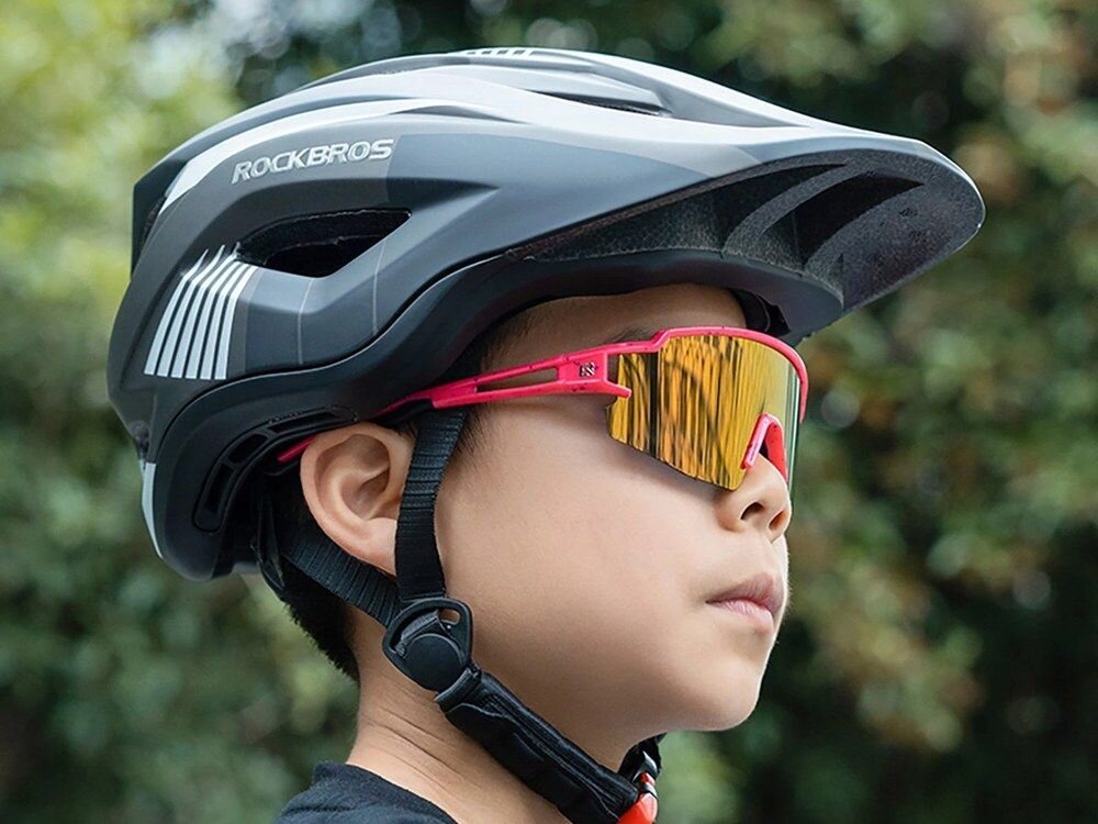 Osoba nosi różowe okulary rowerowe z lustrzanymi soczewkami oraz czarny kask z logo \'ROCKBROS\'. W tle widoczne są rozmyte drzewa.