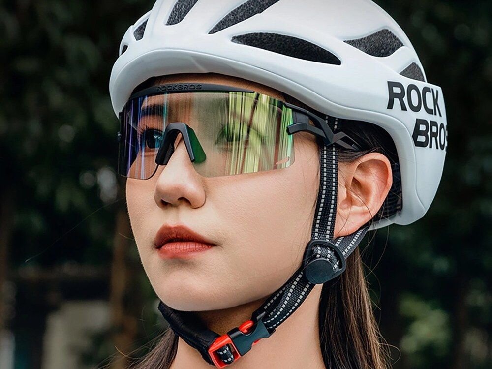 Osoba nosi biały kask z napisem \'ROCKBROS\' oraz okulary przeciwsłoneczne z lustrzanymi szkłami. W tle widoczne są rozmyte drzewa.