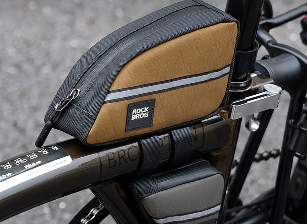 Brązowa torba rowerowa na ramę z czarnymi i szarymi elementami, z widocznym logo \'ROCKBROS\'. Torba jest zamocowana na rowerze, posiada zamek błyskawiczny i odblaskowy pasek.