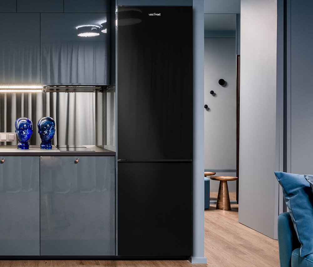 Lodówka VESTFROST VR-FB383-3H0B Na zdjęciu pokazano wnętrze z wysoką, czarną lodówką Vestfrost wkomponowaną w zabudowę kuchenną o grafitowych, połyskujących frontach. Po lewej stronie widać blat z dwoma dekoracyjnymi niebieskimi popiersiami i listwą LED pod szafkami, a po prawej fragment korytarza z drewnianą podłogą i niebieską sofą chłodziarko-zamrażarka wolnostojąca czarne szkło design wysoka jakość komfort użytkowania świeżość wydajność