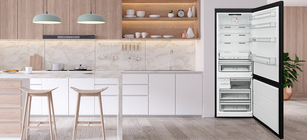 LODÓWKA VESTFROST VR-FB492-3H0B Na zdjęciu przedstawiono jasną, nowoczesną kuchnię z wyspą i dwiema hokerami, utrzymaną w bieli i drewnie z marmurowym wykończeniem ściany. Po prawej stronie stoi czarna lodówka Vestfrost z szeroko otwartymi drzwiami, odsłaniająca przestronne, białe wnętrze z licznymi półkami i szufladami w chłodziarce oraz zamrażarce wygląd styl wnętrze