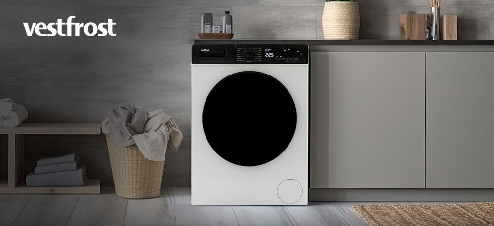 Pralka VESTFROST VW-FW4B1270EU Na zdjęciu znajduje się pralka marki Vestfrost o ciemnym kolorze, wbudowana w szafkę w minimalistycznie urządzonym wnętrzu. Po lewej stronie stoi kosz z jasnymi ręcznikami, a całość aranżacji utrzymana jest w stonowanych barwach i eleganckim stylu