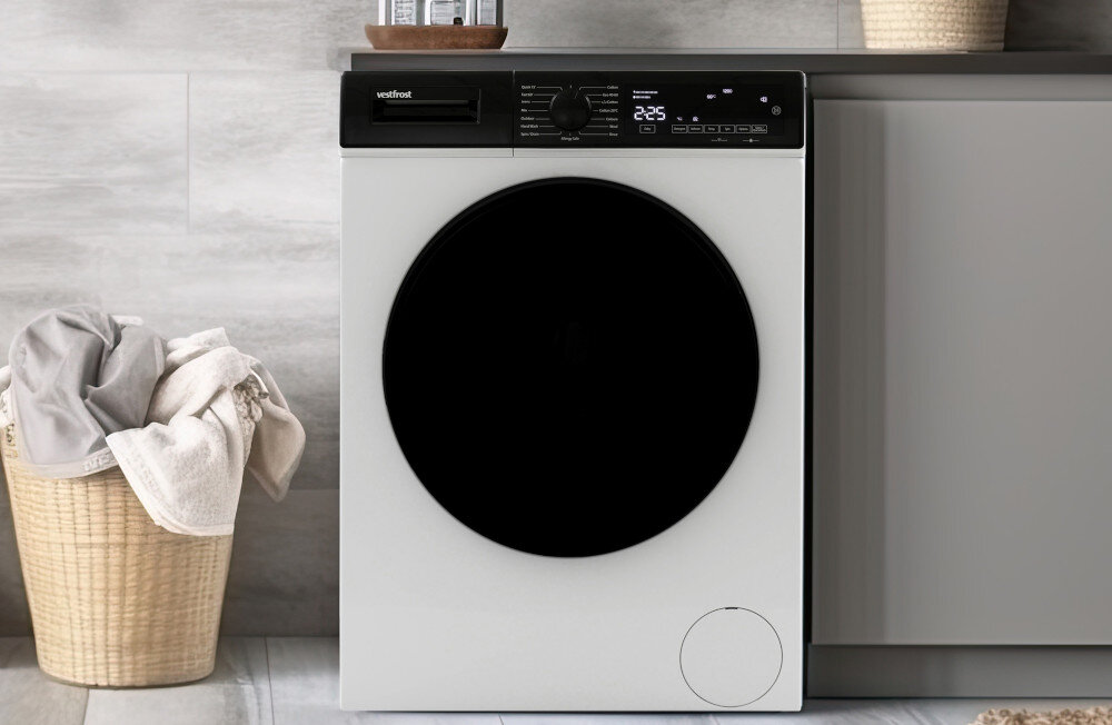 Pralka VESTFROST VW-FW4B1270EU Na zdjęciu znajduje się pralka firmy Vestfrost o czarnej froncie i cyfrowym wyświetlaczu, wkomponowana w zabudowę łazienkową. Po lewej stronie stoi wiklinowy kosz z jasnymi ręcznikami, a cała aranżacja utrzymana jest w jasnych, minimalistycznych kolorach technologia oszczędność funkcjonalność duże drzwi panel sterowania