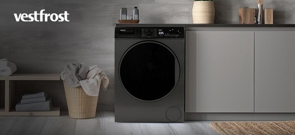 Pralka VESTFROST VW-FD4B1270EU Na zdjęciu znajduje się pralka marki Vestfrost o ciemnym kolorze, wbudowana w szafkę w minimalistycznie urządzonym wnętrzu. Po lewej stronie stoi kosz z jasnymi ręcznikami, a całość aranżacji utrzymana jest w stonowanych barwach i eleganckim stylu