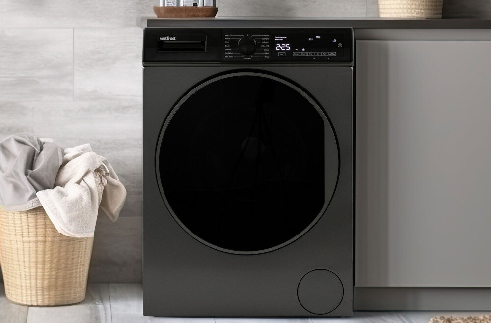 Pralka VESTFROST VW-FD4B1270EU Na zdjęciu znajduje się pralka firmy Vestfrost o czarnej froncie i cyfrowym wyświetlaczu, wkomponowana w zabudowę łazienkową. Po lewej stronie stoi wiklinowy kosz z jasnymi ręcznikami, a cała aranżacja utrzymana jest w jasnych, minimalistycznych kolorach technologia oszczędność funkcjonalność duże drzwi panel sterowania