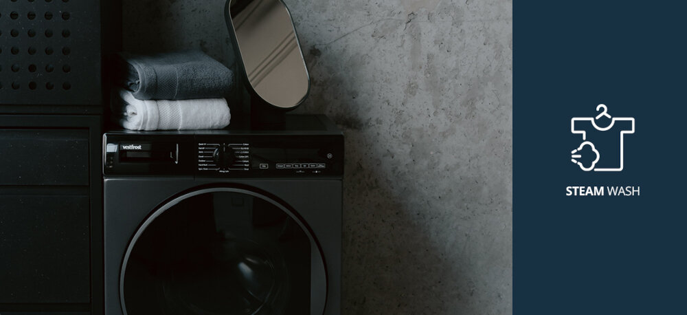Pralka VESTFROST VW-FD4B1270EU Na zdjęciu pokazano pralkę marki Vestfrost z lustrem i złożonymi ręcznikami na górze, ustawioną w stylowym, minimalistycznym wnętrzu. Po prawej stronie grafika informuje o funkcji STEAM WASH (pranie parowe), którą oferuje ten model pralki Steam Wash pranie parowe para wodna mniej zagnieceń higiena prania świeże ubrania alergeny