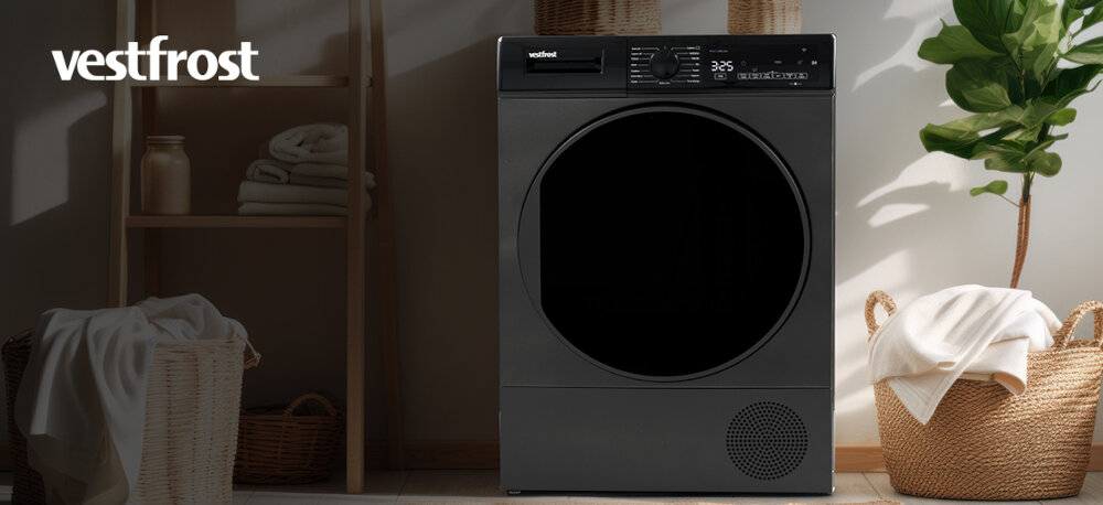 Suszarka VESTFROST VT-FD42HN90EU Na zdjęciu widoczna jest suszarka bębnowa Vestfrost o ciemnym froncie, ustawiona w stylowym, jasnym pomieszczeniu. Po bokach stoją wiklinowe kosze z ręcznikami, a wnętrze jest uporządkowane i dobrze oświetlone naturalnym światłem