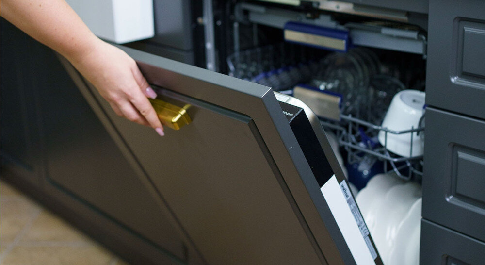 Zmywarka VESTFROST VD-S39B60Q140 Zdjęcie przedstawia częściowo otwartą, zabudowaną zmywarkę z ciemnym frontem meblowym i złotym uchwytem, trzymanym przez dłoń użytkownika. W głębi widać wysunięty kosz z ułożonymi naczyniami, szklankami i miskami, co podkreśla wygodny dostęp do załadunku design inteligentne funkcje cicha praca oszczędność zasobów zabudowa kuchenna estetyka kuchni skuteczne mycie naczyń