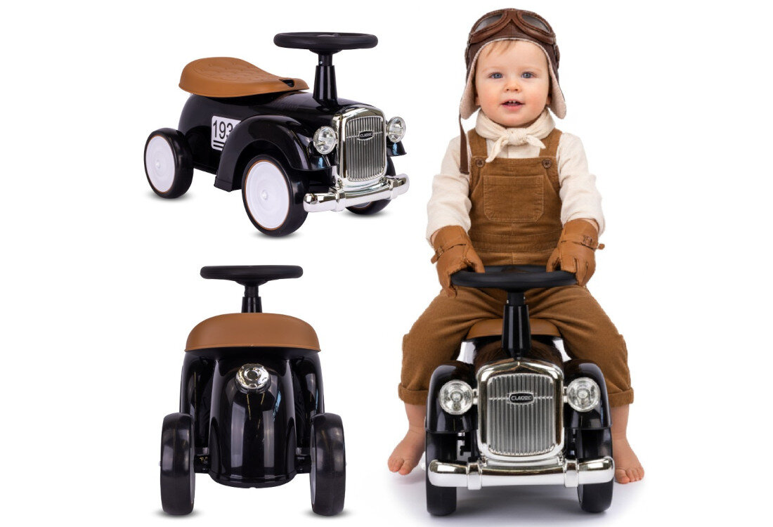Na obrazie widoczny jest czarny jeździk w stylu retro z brązowym siedzeniem i srebrnym grillem. Obok jeździka siedzi dziecko w brązowych ogrodniczkach i czapce lotniczej.
