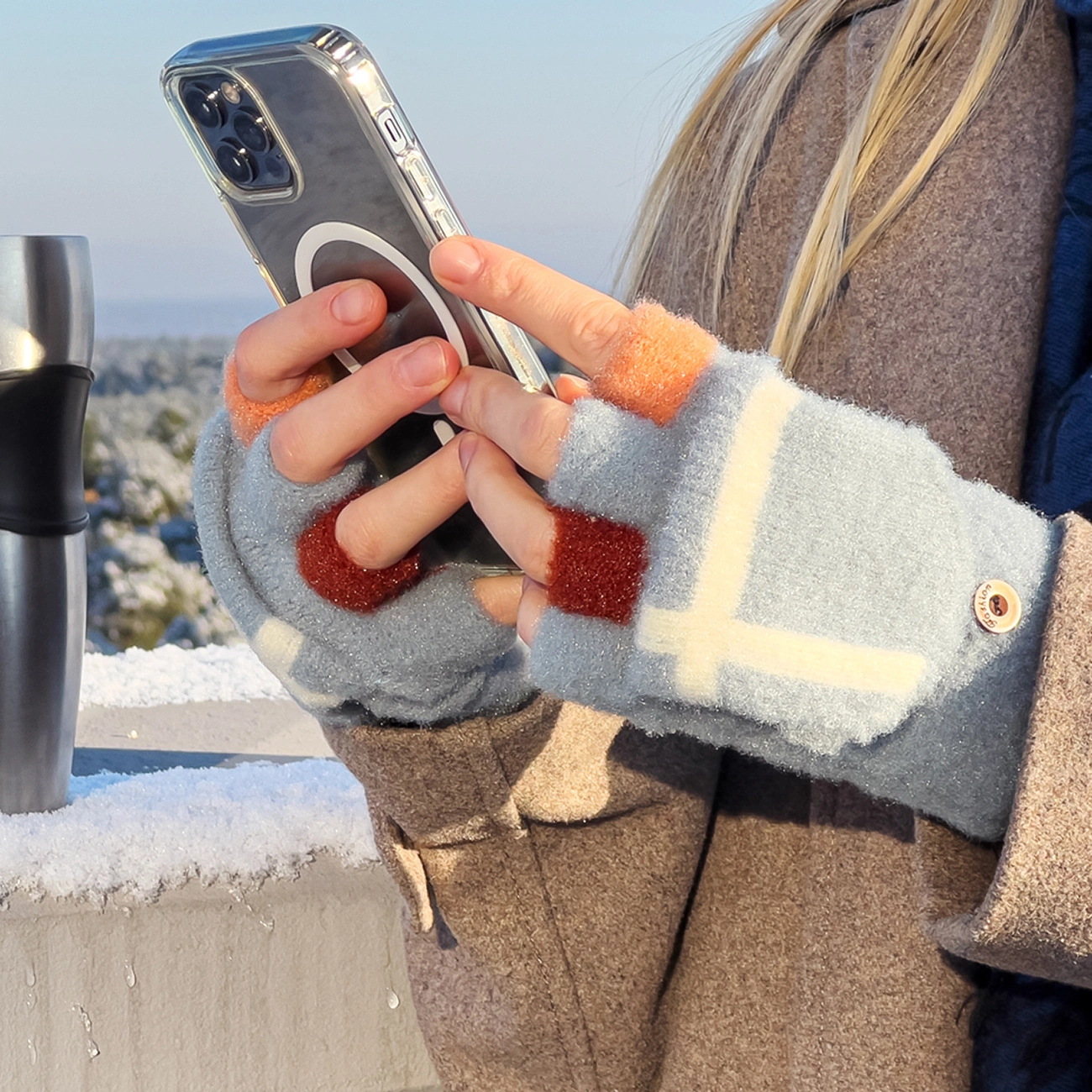 Osoba trzyma telefon w dłoniach ubranych w zimowe rękawiczki z odkrytymi palcami, w kolorze różowym z białymi i brązowymi wzorami. W tle widać zaśnieżony krajobraz i metalowy kubek termiczny na parapecie.