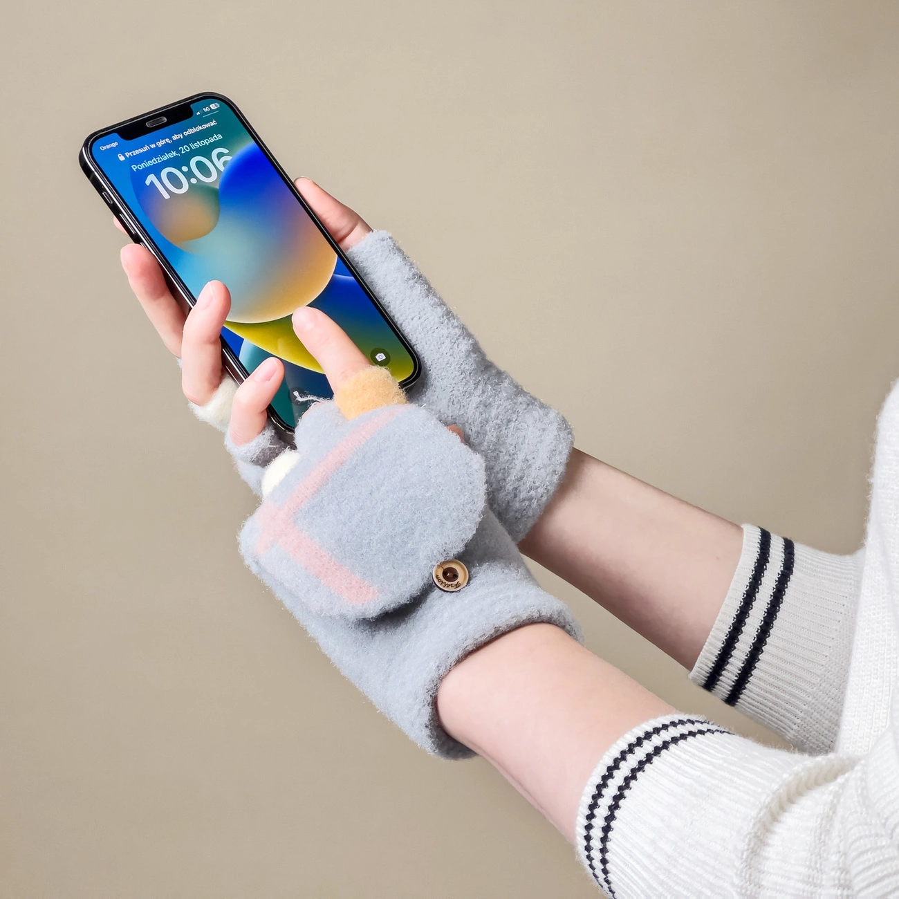 Osoba trzyma smartfon, na którym widoczny jest ekran z godziną 10:06 i tekstem \'Poniedziałek, 16 listopada\'. Na dłoniach ma szare rękawiczki zimowe z odkrytymi palcami i klapką zapinaną na guzik.