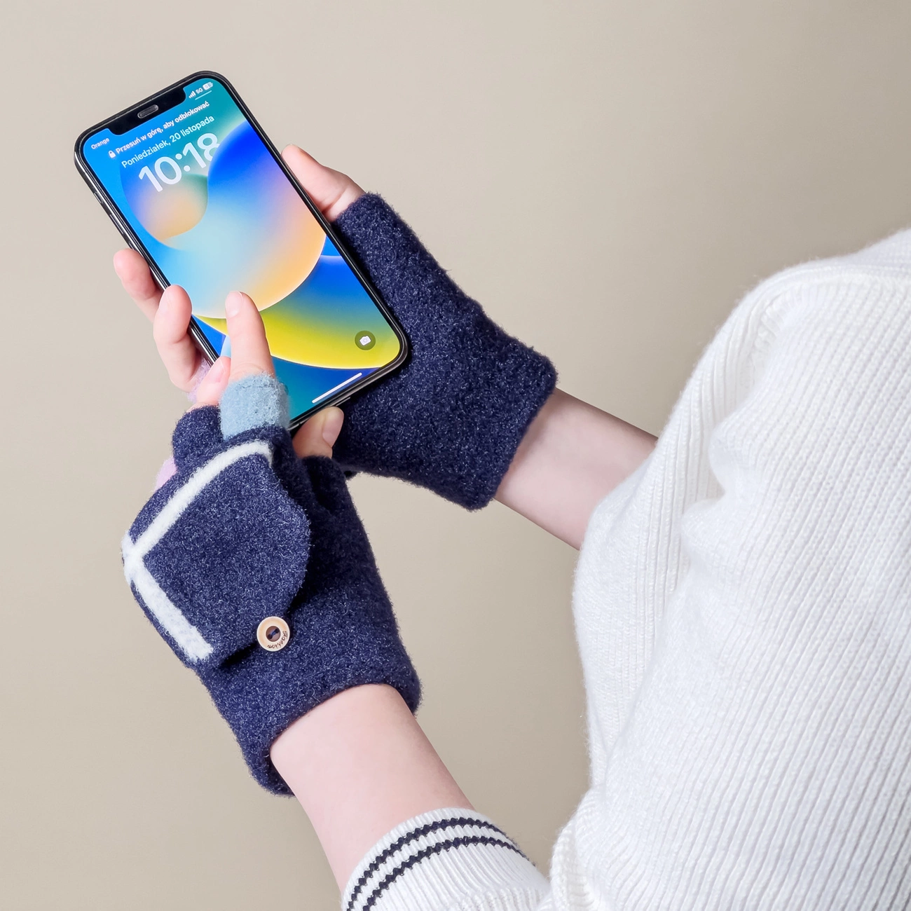 Osoba trzyma smartfon z wyświetlonym ekranem głównym, nosząc granatowe mitenki z białymi i niebieskimi akcentami. Na ekranie telefonu widoczny jest tekst: \'Poniedziałek, 28 listopada\' oraz godzina \'10:18\'.
