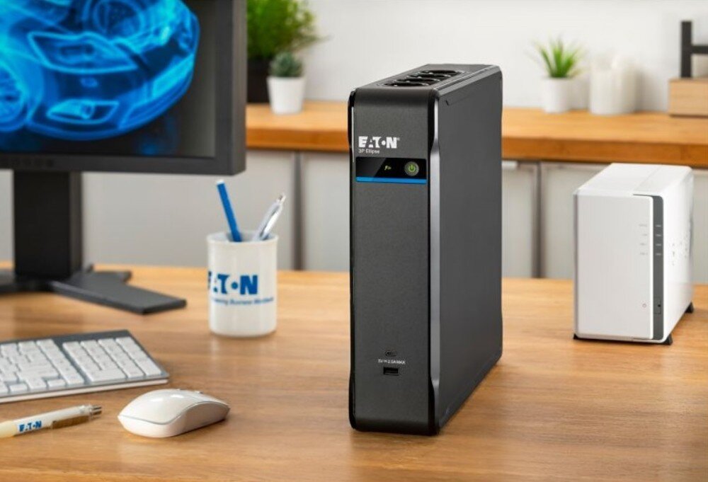 Zasilacz UPS EATON 3P Ellipse Zasilacz awaryjny stojący na drewnianym biurku w biurze, w tle widoczny monitor komputerowy, klawiatura, myszka oraz białe urządzenie sieciowe