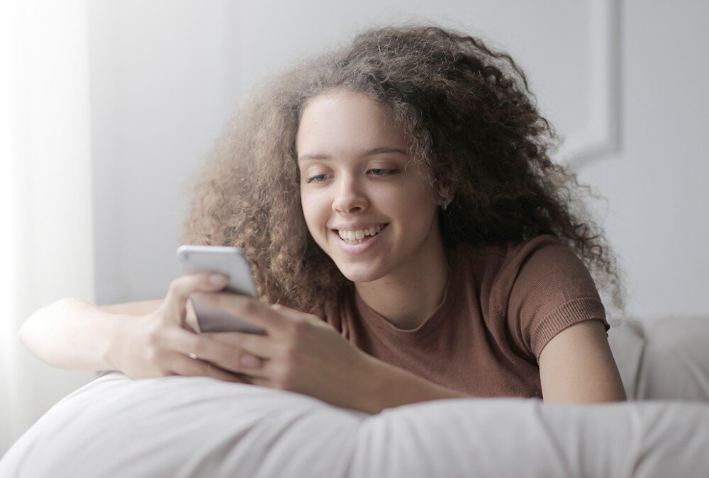 Router MERCUSYS Halo H85X Młoda kobieta z kręconymi włosami leży oparta o poduszkę i trzyma w dłoniach smartfon, patrząc na ekran z uśmiechem