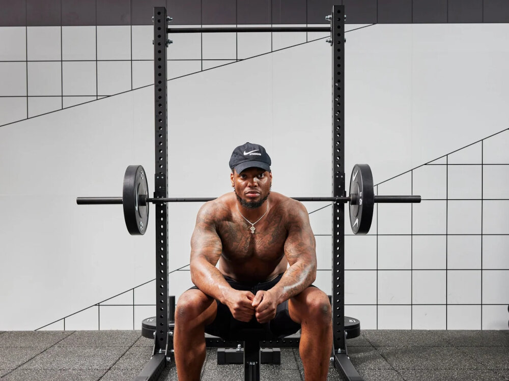 Stojak pod sztangę NIKE Squat Rack sala treningowa osoba siedzi na ławeczce do ćwiczeń za nią stoi stojak wielofunkcyjny trening wszechstrone stanowisko