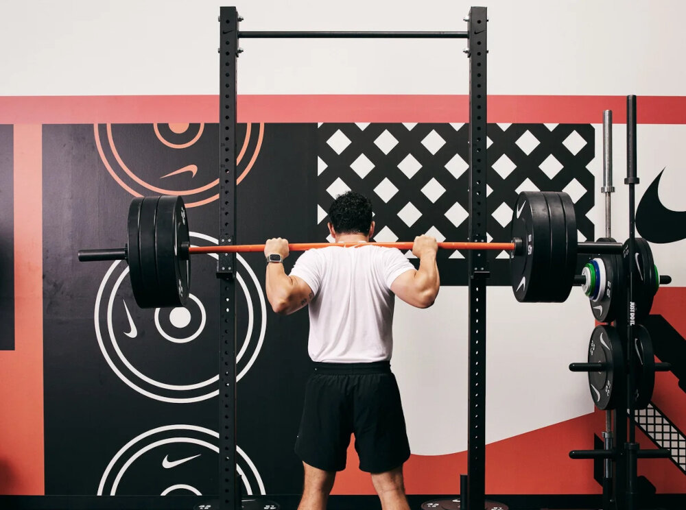 Stojak pod sztangę NIKE Squat Rack sala ćwiczeń osoba ćwiczy ze sztangą przed stojakiem 148 cm długości 126 cm szerokości 236 cm wysokości bez regulowania parametrów przed każdym treningiem Wysoka konstrukcja z drążkiem szeroka podstawa