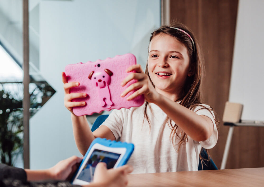 Tablet BLOW Kids Tab 7 V2 7 cali 4/32 GB Wi-Fi Różowy prezentacja urządzenia od tyłu dziewczynka uśmiechająca się i trzymająca tablet w rękach na zajęciach