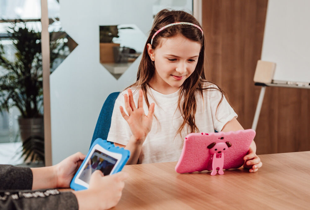 Tablet BLOW Kids Tab 7 V2 7 cali 4/32 GB Wi-Fi Różowy prezentacje urządzenia podczas codziennego korzystania dziewczynka macha ręką w stronę kamery tabletu na zajęciach przedni i tylny aparat 2 MP wideorozmowy zdjęcia nagrywanie filmów funkcjonalność