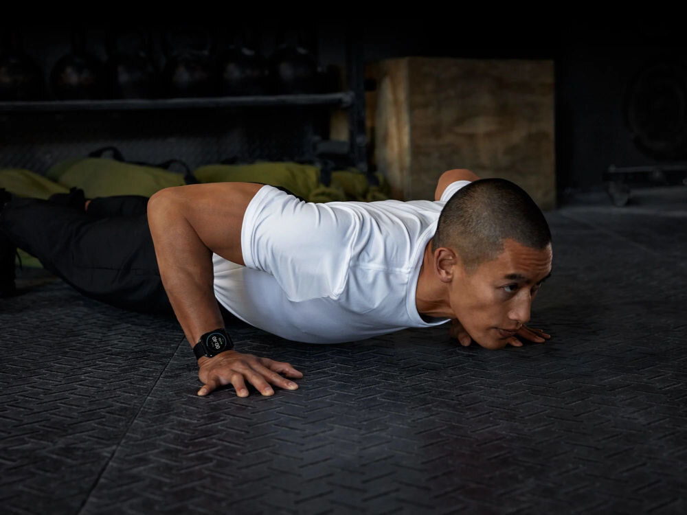 Smartwatch GARMIN Venu X1 Mężczyzna w białej koszulce i ciemnych spodniach robiący pompkę na macie w siłowni/boksie treningowym. Na jego lewym nadgarstku widoczny jest smartwatch. W tle sprzęt sportowy.