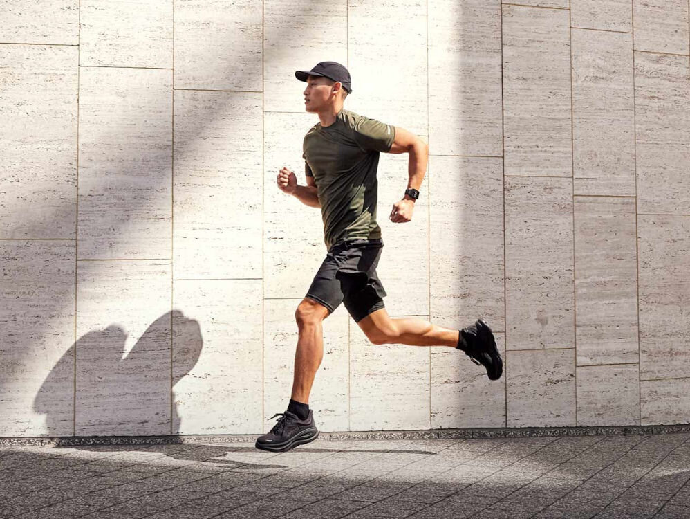 Smartwatch GARMIN Venu X1 Mężczyzna w czapce i sportowym stroju biegnący wzdłuż ściany z białych, teksturowanych płytek, rzucającej długi cień. Ma na sobie smartwatch