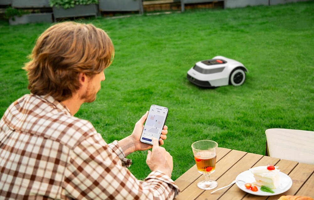 Robot koszący ANTHBOT Pion 1000 trawnik osoba siedzi przy stole trzyma telefon w dłoni w tle robot na trawniku zaawansowany system Cable-TOF tworzenie map 3D w czasie rzeczywistym wykrywa i zgłasza w aplikacji ANTHBOT wszelkie uszkodzenia i przerwy w przewodzie ograniczającym komfort użytkowania pełna kontrola
