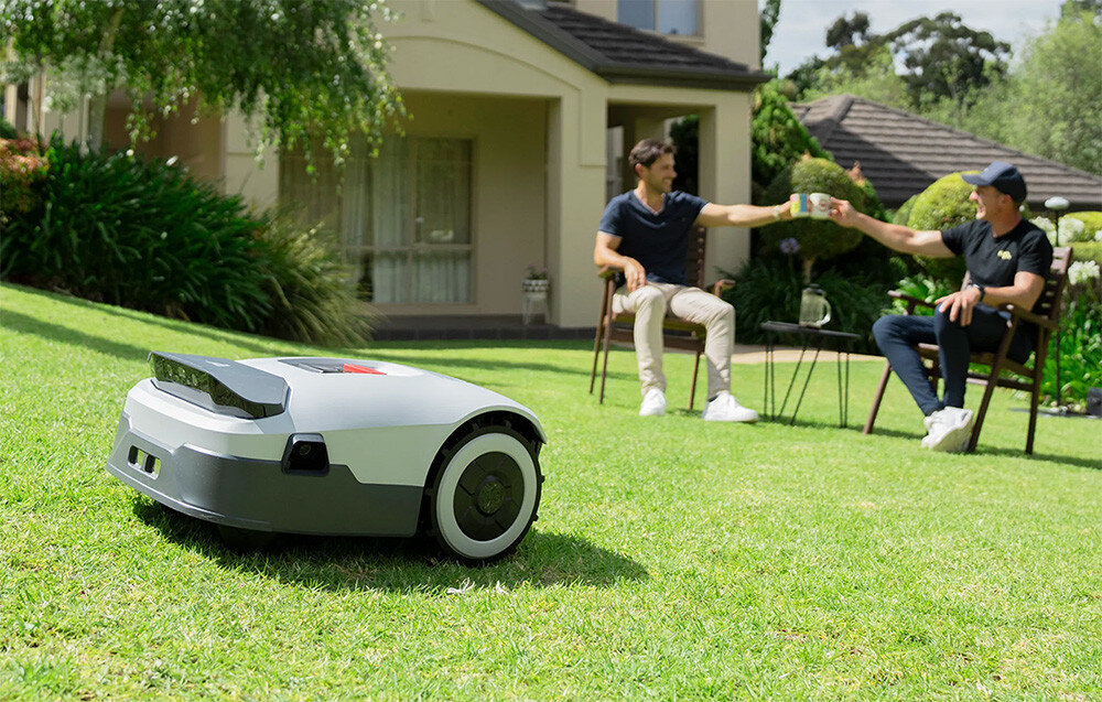 Robot koszący ANTHBOT Genie 3000 trawnik robot kosi trawę w tle dwie osoby siedzą przy stole ogrodowym dalej dom i rośliny inteligentny robot koszący wspierany technologią sztucznej inteligencji aktualizacja OTA regularnie zyskuje nowe funkcje wysoka jakość pracy