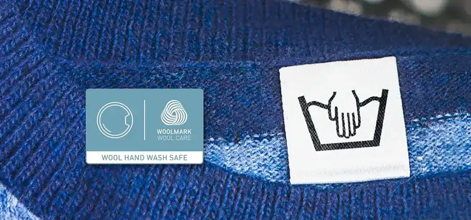 Na obrazie widoczna jest metka z symbolem prania ręcznego oraz niebieska etykieta z logo Woolmark i napisem \'WOOL HAND WASH SAFE\'.