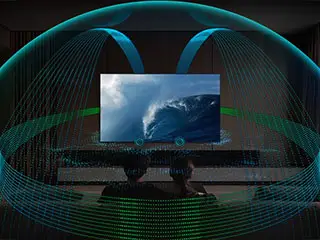 Telewizor wyświetla obraz z falą na ekranie, otoczony wizualizacją dźwięku przestrzennego. W tle widoczne są dwie osoby siedzące przed telewizorem.