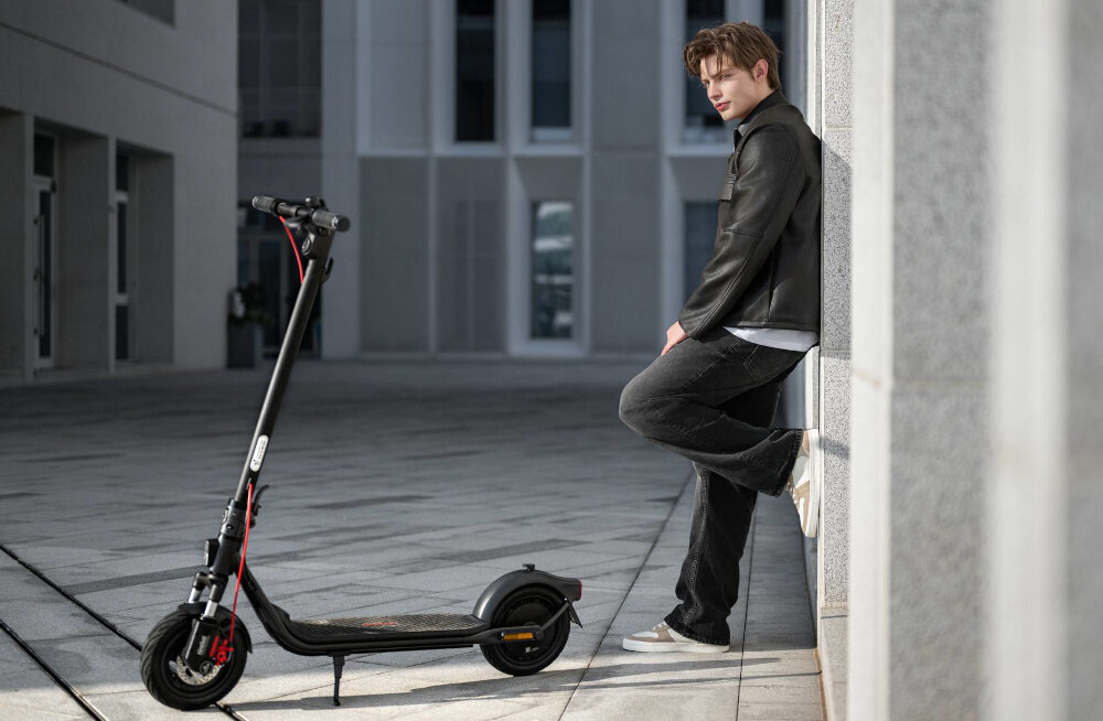 Hulajnoga elektryczna SEGWAY F2 Pro D II 65km 900W 10 cali Czarny miasto budynki osoba stoi oparta o ścianę na pierwszym planie stoi hulajnoga widoczna z boku wnętrzem 10-calowe bezdętkowe opony pneumatyczne typu Tubeless z warstwą żelową automatycznie uszczelniają drobne przebicia jazda bardziej bezpieczna i komfortowa duże amortyzujące uderzenia koła stabilność i przyczepność