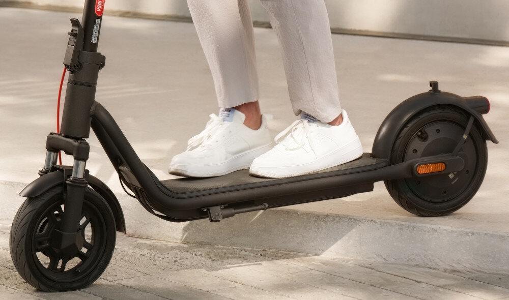 Hulajnoga elektryczna NAVEE V25i Pro 2 20km 300W 10 cali Czarny miasto chodnik zbliżenie na podest osoba jedzie na hulajnodze zjeżdża z krawężnika połączenie stali i aluminium solidność niska waga (17,1 kg) podest o szerokości 152 mm pokryty powłoką antypoślizgową stabilność stopy maksymalne obciążenie do 100 kg minimalizują ryzyko poślizgu