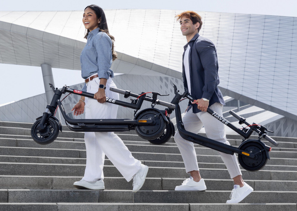 Hulajnoga elektryczna NAVEE V25i Pro 2 20km 300W 10 cali Czarny miasto dwie osoby niosą złożone hulajnogi w dłoniach wchodzą po schodach niebo abstrakcyjny budynek w tle składany mechanizm kierownicę można również złożyć (obrót o 90°) podwójny system składania zmniejsza objętość aż o 66%