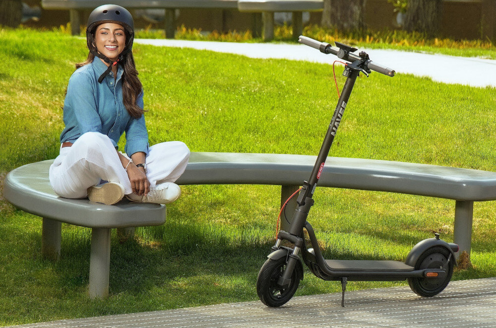 Hulajnoga elektryczna NAVEE V25i Pro 2 20km 300W 10 cali Czarny park trawnik osoba siedzi na ławce obok na oparta na stopce stoi hulajnoga składana stopka wytrzymała nóżka w centralnym punkcie ciężkości zapobiega przypadkowemu przewróceniu się pojazdu