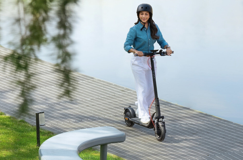 Hulajnoga elektryczna NAVEE V25i Pro 2 20km 300W 10 cali Czarny miasto osoba w kasku jedzie po drodze w tle trawnik woda system KERS odzyskać energię w chwilach hamowania i zamienić ją w dodatkowe kilometry jazdy sprytna technologia hamowanie w realne korzyści i większą swobodę w ruchu