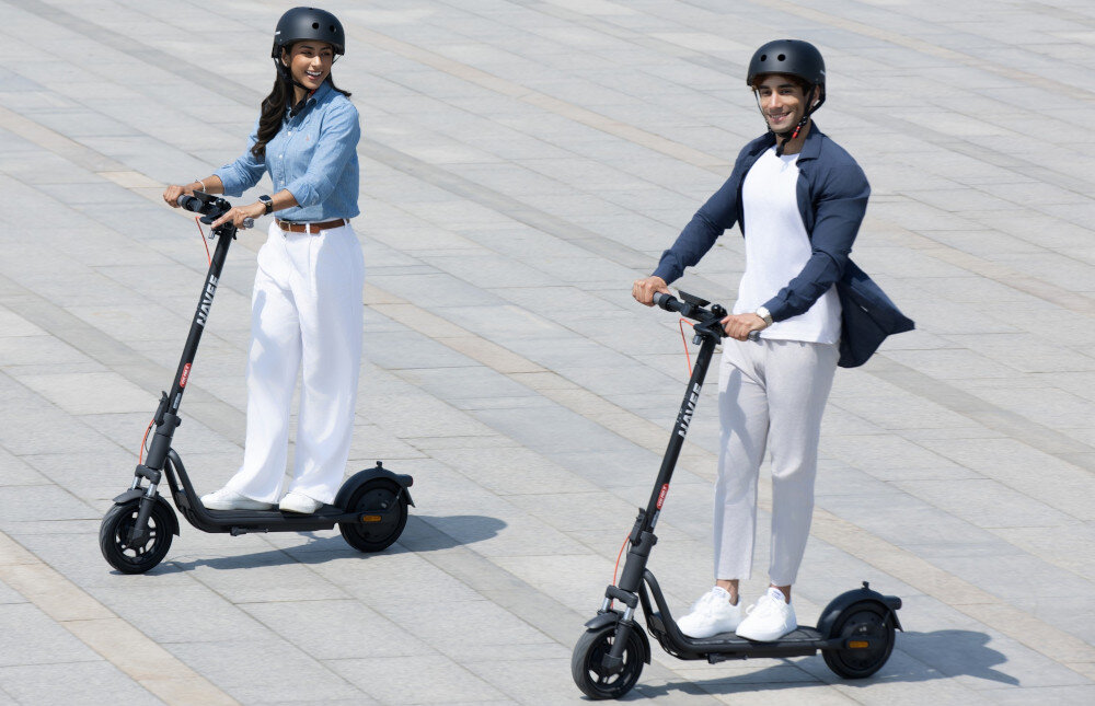 Hulajnoga elektryczna NAVEE V25i Pro 2 20km 300W 10 cali Czarny miasto budynek dwie osoby jadą na hulajnogach chodnik 3 tryby jazdy tryb Spacerowy prędkość do 6 km/h tryb STANDARD prędkość do 15 km/h tryb SPORT prędkość do 20 km/h