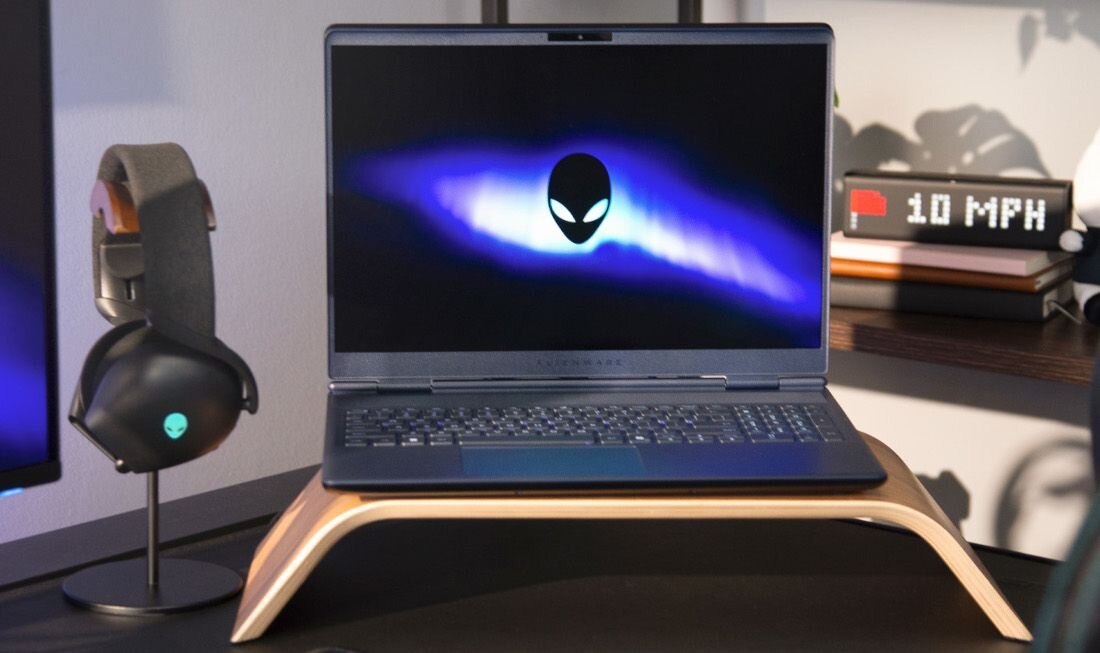 Laptop Alienware na biurku z podświetlanym logo, obok stojak na słuchawki gamingowe i cyfrowy zegar - Intel Core Pamięć RAM DDR5 NVIDIA GeForce RTX Dysk SSD NVMe PCIe Gen4