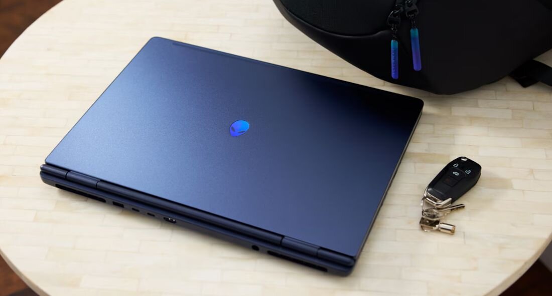 Zamknięty laptop Alienware z podświetlanym logo na okrągłym stole, obok kluczyki i czarny plecak - Opis serii Alienware 16X Aurora