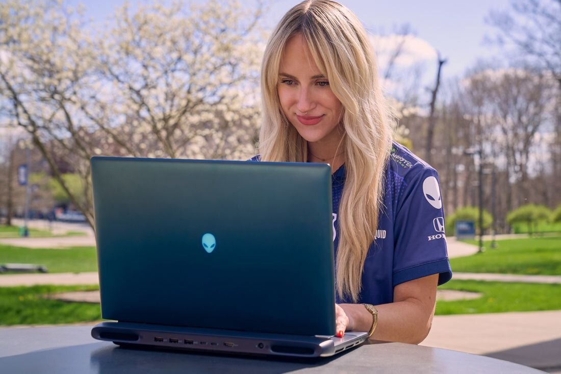  Kobieta w blond włosach w granatowej koszulce Team Liquid z Alienware 16 na świeżym powietrzu, zielony park z drzewami w tle, widok portów z tyłu obudowy laptopa, grafitowa metalowa konstrukcja z logo alien head podświetlanym turkusem, mobilne stanowisko gamingowe outdoor - Standard Wi-Fi 7  