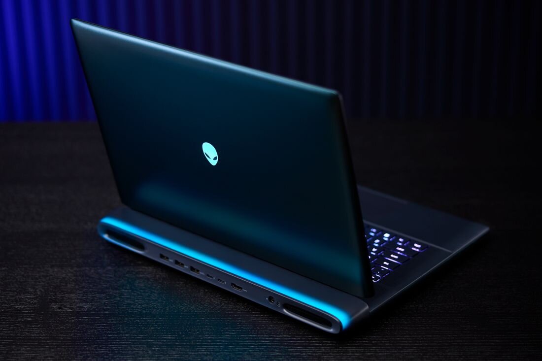 Otwarty laptop Alienware 16 na czarnym drewnianym stole w ciemnym wnętrzu z niebieskimi zasłonami w tle, grafitowa aluminiowa obudowa z podświetlanym turkusowym logo alien head, czarna klawiatura z fioletowym podświetleniem RGB z prawej strony, intensywny niebieski pasek LED na zawiasie z tyłu, widok z boku pokazujący porty tylne i smukłą konstrukcję premium - Bateria 96 Wh Ekologia