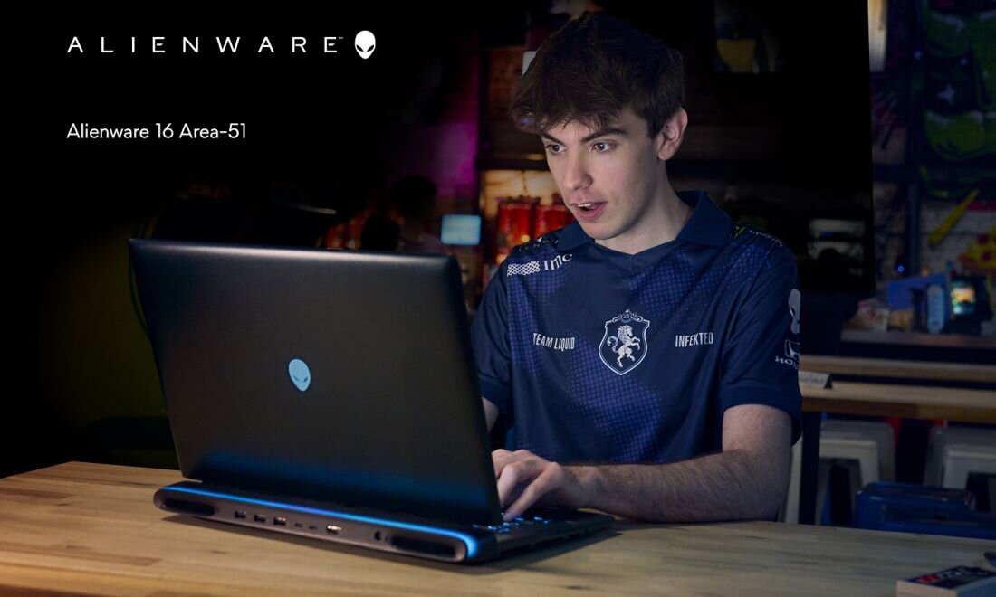  Gracz e-sportowy Team Liquid w granatowej koszulce z logo Infak10 przy laptopie Alienware 16 Area-51 na drewnianym biurku, ekran z boku z podświetlanym logo alien head, niebieskie oświetlenie RGB na krawędzi obudowy, profesjonalne stanowisko gamingowe w pokoju z posterami 
